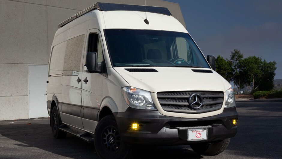 White Mercedes Sprinter van in the daytime in front of building with yellow fog lights on.