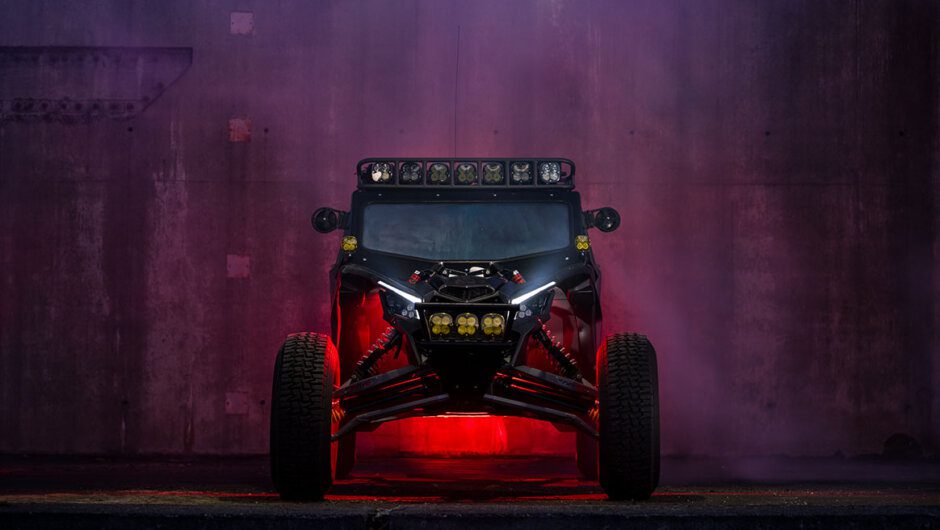 Front view of a custom Can-Am Maverick X3 UTV with an aggressive off-road lighting setup, featuring Baja Designs LED lights and red underglow. The rugged side-by-side is showcased against a moody, smoke-filled urban backdrop, emphasizing its high-performance modifications for night-time adventure riding.